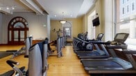 Located at 141 Fourth St E in downtown Saint Paul, Minnesota, this fitness center seamlessly blends historic architectural character with modern exercise amenities. The single-floor facility is set within a building of notable heritage, featuring ornate structural columns with decorative capitals, high ceilings, and an elegant arched entryway with warm wood framing and frosted glass panels that serve as a striking focal point.

The space is divided into distinct workout zones. The cardio area lines the perimeter walls and is outfitted with a row of Matrix treadmills and stationary bikes, each equipped with individual display screens. Large windows along the exterior walls flood this section with natural light and offer views of the surrounding downtown Saint Paul cityscape, with wall-mounted flat-screen TVs providing additional entertainment.

The strength and functional training zone occupies the central and rear portions of the facility, featuring a comprehensive selection of selectorized machines, a cable tower, free weights, dumbbells, medicine balls, and a dedicated stretching area. Rubber flooring covers the weight training areas, transitioning to warm hardwood in the cardio zones. Full-length mirrors are mounted along the walls, enhancing the sense of openness. Recessed and track lighting throughout ensures the entire space is well-illuminated, creating a functional and inviting environment for a complete workout experience.