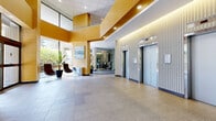 Located at 9500 Erickson Dr in Burnaby, BC, this expansive building lobby presents an impressive and well-appointed hallway that spans a generously sized single floor. The space is defined by its polished terrazzo and large-format tile flooring, which extends throughout the entirety of the area, lending a clean and professional aesthetic. Soaring ceilings clad in warm wood-toned panels rise dramatically overhead, accented by recessed lighting and layered architectural soffits that add visual depth and sophistication.

Floor-to-ceiling glazing runs along multiple exterior-facing walls, flooding the interior with abundant natural light and framing lush greenery and mature trees outside. A pair of stainless steel elevator doors is prominently positioned along one wall, flanked by ribbed column detailing that echoes throughout the space. A glass-enclosed office or amenity suite is accessible directly from the lobby, offering transparency and connectivity.

A tastefully arranged seating area with contemporary chairs and potted plants provides a welcoming focal point, while a central reception-style desk anchors the heart of the lobby. The overall ambiance is modern and refined, balancing open, airy proportions with warm architectural finishes — making a strong and professional first impression for occupants and visitors alike.