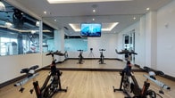 This dedicated spin studio is designed to deliver a focused and energizing cycling experience. The room features a clean, contemporary aesthetic defined by crisp white walls and smooth hardwood-style flooring that adds warmth to the bright, minimal interior. Floor-to-ceiling mirrors line the walls, visually expanding the space and allowing participants to monitor their form throughout a session. A wall-mounted flat-screen television is centrally positioned above the mirror, providing an engaging visual focal point during rides. The ceiling showcases a recessed tray detail with integrated ambient lighting, complemented by additional recessed downlights that illuminate the room evenly. A row of professional-grade stationary cycling bikes is arranged in organized lines, offering ample space between each station for comfortable movement. Glass partitions along one side of the room provide a sense of openness and connectivity to adjacent fitness areas while maintaining the studio's acoustic separation. The overall ambiance is polished and motivating, combining functional design with a sleek, modern finish that supports both individual training and group spin classes.