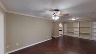 This single-story unit offers a well-organized layout with distinct living, kitchen, bedroom, and bathroom areas. The spacious living room is a central highlight, featuring rich, dark hardwood-style flooring that extends throughout the main areas, crisp white crown molding, and a ceiling fan with integrated lighting. A notable built-in shelving unit with a wine rack accent anchors one wall, adding functional storage and visual interest. An arched passageway connects the living area to the kitchen, which is outfitted with white cabinetry, tile countertops, a tile backsplash, and white appliances including a range, refrigerator, and dishwasher. Light tile flooring completes the kitchen's clean aesthetic. The bedroom is generously proportioned, continuing the dark hardwood-style flooring and crown molding, with a ceiling fan providing comfort and airflow. The bathroom is finished with large-format tile flooring, a tiled tub-shower surround, a white vanity with single-basin sink, and a mirrored medicine cabinet. Textured ceilings and neutral warm-beige walls are consistent throughout, creating a cohesive and practical living environment.