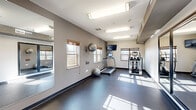 Located at 1290 County Road F W in Arden Hills, Minnesota, this dedicated exercise room offers a well-equipped fitness space designed for efficient, functional workouts. The room features durable dark flooring throughout, providing a practical surface suited for a variety of exercises. Natural light streams in through multiple windows fitted with wood-tone blinds, brightening the space and creating an energizing workout environment, complemented by overhead fluorescent lighting for consistent illumination at all hours.

The fitness area is equipped with a range of cardio and strength training equipment, including a treadmill, elliptical trainer, and a multi-station cable strength machine with an adjustable bench. Stability balls are also available for core and flexibility training. A wall-mounted flat-screen TV adds an entertainment option during workouts. Full-length mirrors along one wall visually expand the space and allow users to monitor their form during exercise.

A convenient wall-mounted water fountain and waste receptacle are positioned near the entry door, ensuring easy access to hydration. The neutral gray and white walls contribute to a clean, modern aesthetic. The space is efficiently organized to maximize usability within its compact footprint, making it a practical and complete fitness facility for residents at this Arden Hills location.