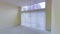 This single-floor space is designed with a clean, minimalist aesthetic that emphasizes openness and natural light. The main area features an expansive wall of floor-to-ceiling windows fitted with white vertical blinds, allowing for flexible light control while maintaining a bright, airy atmosphere. Above the primary window wall, a continuous clerestory window band runs the full width of the room, further enhancing the flow of natural light and providing elevated views of the surrounding urban environment, including neighboring buildings visible from the interior.

The walls are finished in a soft, neutral yellow-white tone that complements the light-colored wall-to-wall carpeting underfoot, contributing to the overall sense of warmth and spaciousness. The ceiling is white and features recessed ventilation grilles, maintaining a streamlined, uncluttered appearance. The absence of furniture and décor highlights the versatility of the space, making it well-suited for a variety of uses. The combination of large glazed openings, neutral finishes, and clean lines gives the space a functional yet inviting character with a distinctly contemporary feel.