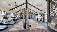Located at 7295 Ascent View in Colorado Springs, CO, this well-appointed fitness center offers a generously sized, single-floor layout designed to accommodate a full range of workout activities. The space is defined by its striking vaulted ceiling accented with dark, rustic wood beams that run the length of the room, lending a warm yet contemporary aesthetic. Recessed lighting, pendant fixtures, and ceiling fans are evenly distributed throughout, ensuring excellent illumination and air circulation. Large windows along the perimeter walls flood the interior with natural light and provide views of the surrounding Colorado landscape. The flooring features a combination of textured, patterned carpet tiles and hard-surface sections that delineate different workout zones. The cardio area is equipped with multiple treadmills, ellipticals, and stationary bikes, each outfitted with individual screens. The strength training zone houses a variety of cable machines, selectorized equipment, and free weight stations, including dumbbell racks paired with a large wall-mounted mirror. A decorative geometric accent wall near the entrance adds a refined design element. Wall-mounted televisions are positioned throughout the space for entertainment. A water fountain station is also available, rounding out this comprehensive, thoughtfully designed fitness facility.