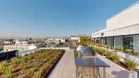This expansive rooftop terrace offers a well-appointed outdoor retreat with a diverse range of amenities set against breathtaking panoramic views of the Los Angeles cityscape, palm tree-lined streets, and distant mountain ranges. The terrace is thoughtfully divided into multiple functional zones, each designed to maximize comfort and outdoor enjoyment.

Lush, landscaped planting beds filled with ornamental grasses, succulents, small trees, and ground cover run throughout the space, adding a vibrant green aesthetic to the clean modern design. Large-format porcelain tile paving provides a sleek, continuous surface across the entire terrace.

A dedicated dining area features a generously sized wooden communal table with colorful seating, perfect for outdoor gatherings. An outdoor kitchen station equipped with a stainless steel grill and prep counter supports al fresco cooking and entertaining. A lounge zone offers reclining chairs, a modern fire pit, and sculptural white planters arranged around a turf lawn area, creating a relaxed, resort-like atmosphere. A ping pong table adds a recreational element to the space.

Glass panel railings line the perimeter, preserving unobstructed sightlines across the surrounding urban landscape. The overall design balances contemporary architecture with natural elements, resulting in a vibrant and versatile outdoor destination.