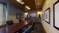 Located at 65 N Madison Ave in Pasadena, California, this single-floor conference room is designed to support professional meetings in a refined and well-appointed environment. The space centers around a polished oval conference table finished in rich mahogany tones, surrounded by ergonomic, high-back leather executive chairs that provide ample seating for a full team.

The room's architectural design features a layered ceiling with recessed lighting panels that cast warm, focused illumination across the table and surrounding area. A large flat-panel display is prominently mounted on one wall within a gold-accented niche, serving as the room's primary presentation focal point. Flanking the screen, decorative tall vases add a curated aesthetic touch, while framed architectural and cityscape photography adorns the surrounding walls, contributing to the room's polished character.

A built-in bench with upholstered cushioning is integrated beneath the display wall, offering additional casual seating. Natural light filters into the room through windows fitted with wood-slat blinds and sheer curtains, providing adjustable brightness. A tall potted plant near the window introduces a natural element to the otherwise formal atmosphere. The dark carpeted flooring and muted gray-green walls complete the room's composed, executive ambiance.
