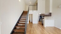 Located at 390 E Main St in Hillsboro, Oregon, this two-story loft-style unit offers a striking open-concept design defined by soaring double-height ceilings that amplify the sense of space throughout the main floor. Light hardwood flooring runs seamlessly across both levels, complementing the bright, neutral palette of the crisp white walls.

The main floor serves as the primary living area, featuring a compact but well-appointed kitchen with dark espresso cabinetry, stainless steel appliances including a range, microwave, and refrigerator, and a white peninsula countertop that provides additional prep and dining space. A wall-mounted mini-split HVAC unit ensures year-round climate control. Large windows allow generous natural light to pour in, casting warm patterns across the open living area.

A sleek open-riser staircase with a bold black steel frame and cable-style railing becomes a central architectural feature, leading up to the mezzanine loft on the second floor. This upper-level space offers a flexible area suited for a sleeping or workspace, with windows providing additional natural light and views to the exterior.

The bathroom is finished with a dark wood vanity, a white countertop with an undermount sink, wood-toned flooring, and accessible grab bars. A hallway and closet complete the layout, providing practical storage and circulation throughout this efficiently designed, modern urban home.