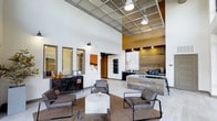 This single-story space makes a strong first impression with its dramatic ceiling height, which creates an open, airy atmosphere throughout the interior. The ceiling features an industrial-style grid panel design with integrated pendant lighting, adding a contemporary edge to the overall aesthetic.

The reception area is a focal point, clad in reclaimed wood paneling that introduces warmth and texture against the crisp white walls. A built-in counter with workstations and display shelving serves as the functional hub of this zone, accented by track lighting that highlights the natural wood tones. An accent wall painted in a rich amber-orange hue adds a bold splash of color, complementing the earthy materials used throughout.

Adjacent to the reception desk is a comfortable lounge seating area, furnished with plush upholstered chairs arranged around a round white coffee table, all anchored by a neutral area rug. Wall-mounted artwork echoes the warm orange and brown color palette, reinforcing the cohesive design theme.

A black industrial-style console along one wall provides additional display and storage space. Natural light filters in through windows, brightening the space and enhancing its welcoming ambiance.
