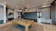 This expansive single-floor amenity space is thoughtfully designed to blend modern industrial aesthetics with inviting comfort. The open-plan layout flows seamlessly between distinct zones, each offering its own character and function. A central bar area anchors the space, featuring a curved countertop with leather bar stools, dark wood beam accents, exposed brick walls, dual mounted TVs, and polished concrete floors that reflect abundant natural light. Adjacent lounge areas provide relaxed seating with tufted leather chairs, sectional sofas, and elegant pendant lighting, all framed by floor-to-ceiling windows that frame views of swaying palm trees and outdoor amenities. A dedicated game room showcases a solid wood pool table beneath geometric pendant fixtures, a shuffleboard table, foosball, and a skee-ball machine, set against a bold striped accent wall. A co-working lounge with long communal tables and industrial-style bar stools sits behind expansive glass walls overlooking a waterfront outdoor deck. A well-equipped kitchenette with stainless steel appliances, a coffee station, and ample counter space supports the entertaining needs of the facility. Throughout, the design incorporates textured accent walls, herringbone carpet inlays, decorative sconces, and curated artwork, creating a cohesive and sophisticated communal environment.