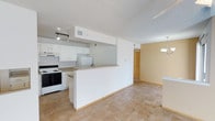 This single-floor space showcases an open-concept layout that connects the kitchen and living area, creating a functional and cohesive environment. The kitchen is equipped with white cabinetry, a white electric range, and a refrigerator, all complemented by a peninsula counter that provides additional workspace and acts as a natural divider between the kitchen and the adjoining living area. Track lighting above the kitchen ensures ample task illumination, while a decorative chandelier adds a touch of warmth near the dining area. The textured popcorn ceiling runs throughout, and the warm beige tile flooring provides a neutral, easy-to-maintain surface across the main living space. Walls are painted in a clean white tone in the kitchen zone, transitioning to a warm yellow hue in the living and dining area, adding visual contrast and depth. A doorway visible in the background leads to additional areas of the unit, hinting at further living space beyond the open common area. Overall, the layout is practical and straightforward, offering a bright, connected environment suited for everyday living.