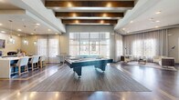 Located at Crescent Pointe Parkway in College Station, Texas, this contemporary single-floor clubhouse offers a well-appointed communal space designed for both relaxation and socializing. The open layout is anchored by polished concrete floors with warm inlay accents, recessed tray ceilings, and abundant natural light filtered through floor-to-ceiling patterned sheer drapes.

The lounge area features a sophisticated seating arrangement of cream swivel chairs and a sleek central coffee table, all set atop a plush area rug. Elegant floor lamps and patterned drapery lend a refined, upscale ambiance to this gathering space.

Adjacent to the lounge, a dedicated game and entertainment zone includes a full-size pool table, a large wall-mounted flat-screen TV set against a textured stacked-stone accent wall, and casual seating with bar-height tables—ideal for informal gatherings.

The kitchen is thoughtfully designed with dark cabinetry, a generous white quartz island, stainless steel appliances, and pendant lighting. A dedicated coffee bar area equipped with a coffee machine, sink, and under-counter refrigerator adds a convenient amenity for daily use. Together, these spaces create a cohesive, modern environment suited for community events and everyday leisure.