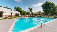 Located at 657 S Lyon St in Santa Ana, California, this single-level outdoor community space offers a well-rounded set of amenities designed for relaxation and recreation. The grounds are anchored by a generously sized swimming pool with stainless steel entry rails and a smooth surrounding deck, accompanied by a series of lounge chairs and a shaded seating area with an umbrella — ideal for unwinding under the Southern California sun. Adjacent to the pool is a circular spa, set into a raised platform on the lush green lawn, providing a dedicated space for relaxation. Mature trees throughout the property offer natural shade and contribute to a park-like atmosphere. A paved courtyard area, flanked by stucco and stone-accented building facades with awning-covered windows, includes outdoor dining furniture beneath a bright umbrella, making it a functional gathering spot. The expansive lawn areas are well-maintained and bordered by iron fencing, adding structure and security to the space. A playground is visible within the fenced grounds, catering to younger residents. The overall ambiance is bright, open, and inviting, reflecting the sunny climate of Orange County.