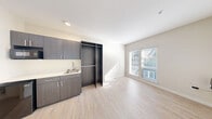 Located at 8500 20th Ave NE in Seattle's Wedgwood neighborhood, this compact yet thoughtfully designed unit offers one bedroom and one bathroom across a single level. The interior features a clean, modern aesthetic with light-toned luxury vinyl plank flooring throughout the main living area, crisp white walls, and recessed lighting that keeps the space feeling bright and open.

The kitchenette area is outfitted with dark charcoal-toned cabinetry, a built-in sink with chrome fixtures, and light countertops — providing a sleek, functional cooking space. An open wardrobe closet with double hanging rods sits adjacent to the kitchen area, offering organized storage without the need for a separate room. Additional closet space with a rod is also integrated into the unit.

The bedroom area receives generous natural light through a set of large windows fitted with horizontal blinds, casting warm, natural light across the pale flooring. The bathroom is clean and utilitarian, featuring neutral flooring, a toilet, and a grab bar, suggesting accessibility-conscious design.

The overall layout is highly efficient, making smart use of every square foot. The minimalist, contemporary finishes — including the dark cabinetry contrast against light walls and floors — give the unit a modern, cohesive character well-suited for urban living in Seattle.