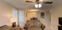 Located at 4223 I-20 Frontage Rd in Vicksburg, Mississippi, this single-floor property offers a functional and comfortable layout designed for everyday living. The open-concept living and dining area serves as the heart of the home, where soft carpeted flooring and neutral-toned walls create a warm, inviting ambiance. A decorative ceiling fan with integrated lighting anchors the living space, while an elegant chandelier illuminates the adjacent dining area, which comfortably accommodates a dining table and chairs. The living room provides ample space for seating and entertainment, with room for a media console and television. A pass-through opening connects the main living area to the kitchen, offering a sense of openness while maintaining a defined separation between spaces. The kitchen features dark cabinetry and is accessible via a dedicated entry from the main living corridor, providing both convenience and practicality. The overall layout is straightforward and efficient, making excellent use of the available footprint. The consistent color palette and understated finishes throughout give the space a cohesive, well-maintained character that is both functional and welcoming.