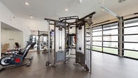 Located in Goodyear, Arizona, this fitness center offers a comprehensive and thoughtfully designed exercise space on a single level. The open-concept layout is bright and inviting, featuring crisp white walls, recessed lighting, and durable rubber flooring suited for high-intensity use. Large glass-panel garage doors line one side of the room, flooding the interior with natural light and providing views of the surrounding outdoor green space — creating an energizing atmosphere that seamlessly connects the indoors with the outside.

The facility is outfitted with a diverse range of professional-grade Life Fitness equipment. A central Life Fitness SYNRGY360 functional training system serves as the anchor of the space, accommodating cable work, TRX suspension training, and battle ropes. Surrounding it is an array of free weights, including a full dumbbell rack, kettlebells, and a multi-station barbell and weight bench setup. Cardio equipment, including treadmills and an elliptical, is positioned near the glass doors to take advantage of the natural light and outdoor views. Additional accessories such as medicine balls, stability balls, and resistance tools are neatly organized on dedicated racks. Full-length mirrors amplify the sense of space, while mounted flat-screen televisions add entertainment options for users. A separate entry door provides convenient, direct access to the facility.