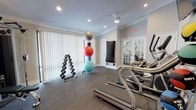 This single-story amenity space is thoughtfully designed to offer residents a comfortable and functional environment for both relaxation and fitness. The main lounge area is anchored by vaulted, beam-accented ceilings that lend an airy, open feel, complemented by large windows with vertical blinds that flood the room with natural light and offer outdoor views. The lounge features a tufted sofa, coffee table, and a wall-mounted television set against a textured stacked-stone accent wall, adding warmth and visual interest. An adjacent kitchenette provides white shaker cabinetry, a mosaic tile backsplash, stainless steel appliances including a microwave and compact refrigerator, and a single-basin sink — well-suited for casual refreshments. The dining area extends naturally from the lounge, featuring high-top bar tables with industrial-style seating beneath the vaulted ceiling, bathed in light from floor-to-ceiling windows. French doors connect the lounge to the fitness center, which is outfitted with rubber flooring, crown molding, a ceiling fan, cardio machines including treadmills, a multi-station cable machine, dumbbell rack, and exercise balls, creating a comprehensive workout facility within an attractive, well-lit setting.