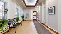 This building's common area consists of a welcoming lobby and an adjoining corridor, together offering a refined and functional entrance experience. The lobby immediately impresses with its signature tray ceiling framed in rich wood molding, complemented by recessed lighting that creates a warm, inviting ambiance. Cream-toned tile flooring extends throughout, accented by a runner mat near the main entrance. White wainscoting and panel molding line the walls, lending a classic, elegant character to the space. A decorative wrought-iron bench and lush potted greenery add warmth and charm, while large windows allow natural light to filter in and provide views of the surroundings. A wooden interior door leads further into the building, and a wall-mounted mailbox unit serves residents conveniently near the main entry. The adjoining corridor transitions to patterned carpet flooring and continues the wood-trim ceiling detail. This hallway is equipped with a stainless steel elevator, a bulletin board, and additional framed artwork along the walls. A hand sanitizer station and directional signage reflect practical amenities integrated seamlessly into the well-maintained space. Together, the lobby and corridor present a polished, professionally managed building entrance.