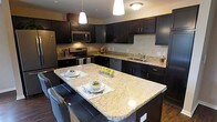 This single-floor residence offers a thoughtfully designed layout that balances modern style with everyday functionality. The open-concept kitchen is a standout feature, showcasing dark espresso cabinetry that contrasts elegantly with light granite countertops. A generously sized center island with pendant lighting overhead provides both additional prep space and casual seating, making it ideal for entertaining. Stainless steel appliances, including a refrigerator, range, and microwave, complete the kitchen's polished, contemporary look. Dark hardwood flooring flows seamlessly from the kitchen into the living area, which is bathed in natural light from multiple large windows offering views of the surrounding neighborhood. The living room provides a comfortable and open gathering space with ample room for seating arrangements. The bedrooms are carpeted for warmth and comfort, each featuring large windows that invite natural light and outdoor views. The bathroom continues the home's refined aesthetic with matching dark cabinetry, granite countertops, a large vanity mirror, and hardwood flooring. Throughout the space, neutral wall tones provide a versatile backdrop that complements the rich, dark finishes, creating a cohesive and inviting atmosphere.