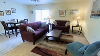 This single-floor space offers a welcoming and functional layout designed for comfortable everyday living. The living area serves as the heart of the home, featuring plush carpeting underfoot and warm, neutral wall tones that create a relaxed, inviting atmosphere. A pair of deep burgundy sofas are arranged around a dark wood coffee table, anchored by a rich, earth-toned area rug that adds texture and warmth to the room. Decorative accent pillows complement the color palette, lending a thoughtful, finished look to the seating arrangement. Natural light filters into the living area through a window dressed with vertical blinds, brightening the space and enhancing its open feel. A ceiling fan overhead provides both comfort and airflow. Tasteful wall art and ambient floor and table lamps contribute to the cozy, lived-in character of the room. A glimpse of a dining area is visible adjacent to the living space, suggesting a practical, open flow between shared living zones. Overall, the space presents a comfortable, no-fuss environment with a warm color scheme and functional design that makes it well-suited for relaxed, day-to-day living.