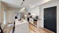 This well-appointed single-floor residence offers a thoughtfully designed layout that blends modern aesthetics with everyday functionality. The open-concept kitchen serves as a centerpiece, featuring crisp white shaker cabinetry extending to the ceiling, a herringbone tile backsplash, stainless steel appliances, and a striking quartz waterfall island illuminated by a cluster of globe pendant lights. Light wood flooring runs throughout, adding warmth to the clean, contemporary palette.

The living area is bright and inviting, benefiting from a large multi-pane window that draws in natural light and provides views of the surrounding area. Neutral-toned walls create a versatile backdrop for the comfortable, mid-century modern furnishings.

The bedrooms are carpeted for comfort, each equipped with ceiling fans and windows that allow for natural ventilation and light. Several bedrooms feature dedicated walk-in closets with built-in shelving and hanging storage, offering generous organization space. The neutral gray walls and clean lines give each room a calm, restful atmosphere.

Two bathrooms serve the residence — one with a glass-enclosed walk-in shower and a white vanity with dark countertop, and a second with a tiled shower-tub combination. Both bathrooms maintain a cohesive, clean design with framed mirrors and tile finishes throughout.