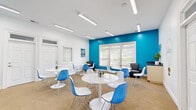This single-floor workspace is a well-appointed, open-plan room designed to foster collaboration and creativity. The interior makes a strong visual statement with a bold teal accent wall that contrasts sharply against crisp white walls and ceiling, creating an energetic yet professional atmosphere. Linear LED light fixtures are mounted across the ceiling, providing consistent, bright illumination throughout the space, complemented by natural light streaming through multiple windows fitted with white horizontal blinds.

The room is furnished with several round white pedestal tables, each surrounded by a mix of white and blue modern shell chairs with chrome wire bases — a cohesive color scheme that ties seamlessly into the overall design. The flooring is a neutral, textured carpet that adds warmth and helps manage acoustics.

A mounted whiteboard on the teal wall serves as a focal point for presentations or brainstorming sessions, while a small workstation with office equipment is positioned nearby. A decorative light bulb wire art piece labeled "ideas" reinforces the creative intent of the space. Multiple entry doors with transom windows are positioned along the walls, offering convenient access and adding to the room's airy, open feel. This space is well-suited for small group meetings, training sessions, or collaborative work environments.