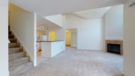 This two-story residence is defined by its dramatic vaulted ceilings in the main living area, which create a sense of openness and volume rarely found in similarly scaled homes. The living room is finished with neutral wall-to-wall carpeting and flows seamlessly into the dining and kitchen area, separated by a white breakfast bar with a dark wood countertop. The kitchen is efficiently laid out in a galley style, featuring light wood cabinetry, dark countertops, tile flooring, and a full suite of white appliances including an electric range, over-the-range microwave, and refrigerator. A dedicated laundry room houses a full-size top-loading washer and dryer. The bedrooms are carpeted and bright, each fitted with ceiling fans and ample closet space. One bedroom includes a large window with vertical blinds and a baseboard heater. The bathrooms are finished with light tile flooring, and one features an extended vanity with light wood cabinetry, a stone-toned countertop, and a soaking tub with a white surround. A utility access panel is also present in one of the lower-level rooms, adding practical functionality to the layout. Throughout, a consistent white and neutral palette gives the space a clean, airy character.