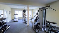 This fitness center offers a bright and functional workout environment designed to accommodate a variety of exercise routines. The space is finished with durable carpet flooring in a neutral tone, providing comfort and stability underfoot during workouts. Light yellow walls and recessed lighting, complemented by a ceiling fan, keep the atmosphere well-lit and comfortable. Multiple large, white-framed windows line the walls, allowing abundant natural light to fill the room and create an open, airy feel. A wall-mounted television is positioned for easy viewing during cardio sessions. The cardio area features multiple treadmills and an elliptical trainer, while the strength training section is equipped with a multi-function cable machine and a weight bench, offering a comprehensive range of resistance exercises. A structural column with a clean, white finish is integrated into the layout without impeding the usable floor space. The overall design is clean and practical, with a straightforward layout that ensures ease of movement between equipment. This fitness center provides all the essential amenities needed for a complete and effective workout experience.