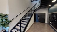 This lobby space presents a well-designed, contemporary environment that combines functional amenities with modern industrial aesthetics. The standout architectural feature is an open staircase with sleek black metal railings and glass panel balustrades, rising through a dramatic double-height volume that allows natural light to filter down from upper-level windows, creating an airy, spacious atmosphere.

The polished concrete floors extend throughout the lobby, reinforcing the industrial character of the space. The walls are finished in neutral tones, complemented by deep teal accent walls that add a sophisticated pop of color. Overhead, recessed and pendant lighting provides warm, focused illumination throughout the corridor.

A comprehensive mail and package management system lines one wall, featuring an extensive bank of individual black mailboxes alongside a Luxer One smart package locker system, offering residents a secure and convenient solution for parcel retrieval. A small marble-topped console table with a waste bin nearby provides a practical spot for sorting mail.

Potted fiddle-leaf fig trees are positioned near the staircase, introducing organic texture and greenery that softens the industrial elements. Overall, the lobby conveys a clean, modern sensibility with thoughtful attention to both design and resident functionality.