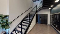This lobby space presents a well-designed, contemporary environment that combines functional amenities with modern industrial aesthetics. The standout architectural feature is an open staircase with sleek black metal railings and glass panel balustrades, rising through a dramatic double-height volume that allows natural light to filter down from upper-level windows, creating an airy, spacious atmosphere.

The polished concrete floors extend throughout the lobby, reinforcing the industrial character of the space. The walls are finished in neutral tones, complemented by deep teal accent walls that add a sophisticated pop of color. Overhead, recessed and pendant lighting provides warm, focused illumination throughout the corridor.

A comprehensive mail and package management system lines one wall, featuring an extensive bank of individual black mailboxes alongside a Luxer One smart package locker system, offering residents a secure and convenient solution for parcel retrieval. A small marble-topped console table with a waste bin nearby provides a practical spot for sorting mail.

Potted fiddle-leaf fig trees are positioned near the staircase, introducing organic texture and greenery that softens the industrial elements. Overall, the lobby conveys a clean, modern sensibility with thoughtful attention to both design and resident functionality.