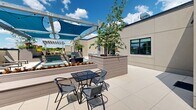 Located at 600 North Avenue in New Rochelle, NY, this rooftop terrace offers a well-appointed outdoor retreat designed for both relaxation and social gathering. The space is anchored by a striking pergola structure draped with vibrant teal shade sails that provide comfortable relief from direct sunlight while string lights overhead add an inviting ambiance for evening use. The large-format tile flooring, rendered in a clean mix of white and teal, lends the terrace a modern, polished aesthetic. Generously sized raised planter beds constructed with wood-tone composite panels are integrated throughout the space, featuring ornamental trees, flowering plants, and greenery that bring a lush, garden-like quality to the rooftop setting. Multiple seating configurations cater to varied uses: cushioned wicker lounge chairs and sofas create intimate conversation areas, while a metal dining table with coordinating chairs provides a spot for al fresco meals. Sun loungers positioned toward the open sky invite residents to unwind and take in the expansive views of the surrounding New Rochelle landscape. An outdoor kitchen station with a built-in grill adds functional amenity to the space. Glass double doors connect the terrace back to the building interior, ensuring seamless indoor-outdoor flow throughout.