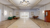 Located at 1707 Main St in La Crosse, Wisconsin, this thoughtfully designed single-floor amenity space offers an impressive range of functional areas finished with a warm, modern aesthetic. The grand entrance lobby sets a welcoming tone with its herringbone-patterned hardwood floors, ornate coffered ceiling, crown molding, and a statement brass chandelier, creating an immediately elegant first impression. Adjacent to the entry, an expansive open living area continues the herringbone flooring and features exposed architectural beams, ample room for lounge seating, and a dining arrangement accented by curated artwork. A dedicated lounge area offers a tufted leather sofa, large wall-mounted display, and sage-green accent walls alongside light wood flooring, providing a comfortable retreat. The kitchen and living area combines warm wood cabinetry, open shelving, a stainless steel French-door refrigerator, and a central island with clean white countertops. A smaller carpeted meeting room with recessed panel ceiling and soft lighting offers a quiet, intimate setting. The fitness center is generously appointed, housing a full cable machine, free weights, mirrors, treadmills, ellipticals, and a stationary bike across two dedicated exercise rooms with carpet flooring and bright panel lighting throughout.