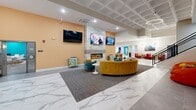 Located at 1000 Elm St in Manchester, NH, this single-floor amenity space offers an impressive and generously sized layout designed for community living and recreation. The interiors are defined by a bold, contemporary aesthetic — featuring vivid teal accent walls, warm orange ceiling insets, and a mix of marble-look tile and wood-plank flooring throughout.

The central lounge area serves as the heart of the space, anchored by a sleek linear fireplace clad in marble-look tile, flanked by multiple wall-mounted TVs, and furnished with curved velvet sofas in rich mustard and teal tones. Adjacent to this is a well-appointed game room offering a billiards table, shuffleboard table, air hockey table, foosball, and arcade games — all set beneath stylish pendant and recessed lighting.

A dedicated co-working and dining area features a long white communal table with upholstered navy and gold bar-height chairs, globe pendant fixtures, and teal accent walls adorned with local photography. A glass-enclosed library nook with custom shelving provides a quiet retreat.

The fitness center is visible through a large interior window, equipped with stationary bikes and exercise equipment beneath circular pendant lighting. A snack and vending area rounds out the offerings, ensuring convenience for all users. The overall ambiance is energetic yet refined, making this a dynamic and welcoming community amenity floor.