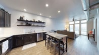 This single-floor space showcases an open-concept layout that effortlessly blends a functional wet bar area with a comfortable living space. The wet bar is a standout feature, complete with dark espresso cabinetry, a stainless steel sink, a dishwasher, and a white subway tile backsplash. Open floating shelves above the bar display decorative items and accessories, adding personality to the space. Recessed lighting illuminates the area from above, highlighting the clean white walls and smooth ceiling.

A high-top bar table with industrial-style metal stools serves as a natural dividing element between the bar and the living area, offering additional seating and casual dining options. The living space beyond features a neutral-toned sofa, a small accent table, and a ceiling fan, creating a relaxed atmosphere. Large windows along the far wall allow abundant natural light to flood the room, contributing to the bright and airy feel. Warm wood-look flooring runs throughout, unifying the entire space with a cohesive and contemporary aesthetic.