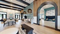 The Tortola clubhouse is a generously proportioned single-floor community space that seamlessly integrates a living room, dining area, and kitchen within a sophisticated coastal-contemporary design. The entry foyer features a striking moss wall backdrop, warm wood slat paneling, exposed dark ceiling beams, and light-toned hardwood floors that carry throughout the space.

The living area is anchored by a herringbone-tiled fireplace flanked by floor-to-ceiling black-framed glass doors, flooding the room with natural light and outdoor views of palm-lined surroundings. Built-in shelving, a wall-mounted TV, and curated furnishings add comfort and function.

The dining zone showcases a dramatic teal mosaic tile feature wall, oversized woven pendant lights, and communal tables with stylish seating. Adjacent is a coffee station with warm wood cabinetry. The kitchen is defined by its bold black-and-white geometric tile flooring, a long white quartz island with bar seating, natural wood lower cabinets, and sculptural gold pendant lighting.

A separate game room features a billiard table, wall-mounted TV, and a teal-painted ceiling accent. Elegant arched doorways connect the zones, creating a cohesive, resort-inspired atmosphere throughout.