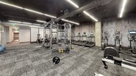 This contemporary fitness center is designed to deliver a comprehensive workout experience across a single, thoughtfully organized floor. The main gym floor exudes an industrial-modern aesthetic, featuring exposed ductwork along a dark ceiling, textured accent walls, and sleek linear LED lighting that illuminates the space with a focused, energetic ambiance. The floor is finished with durable, patterned carpet tile throughout the workout areas.

The cardio zone is equipped with a row of Life Fitness treadmills, each fitted with individual screens, alongside Cybex Arc Trainers and additional elliptical machines. The strength training area features Life Fitness Synrgy functional training rigs with TRX suspension straps and cable attachments, free weights including barbells and bumper plates, medicine balls, and a dedicated dumbbell rack. Cybex benches and weight machines round out the strength offerings.

A glass-walled cycling studio provides a separate, focused environment for spin classes, housing several indoor cycling bikes and offering visibility into the broader gym floor. Adjacent to the main workout area, a bright reception and check-in space features tile flooring, a front desk, and a refreshment station, creating a welcoming entry point. Rowing machines and additional functional training equipment occupy open floor space, ensuring versatile training options throughout the facility.