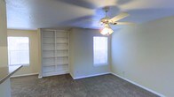 This single-story space presents a well-appointed room characterized by its neutral, understated aesthetic. The room features wall-to-wall carpeting in a warm beige tone, providing a comfortable and cohesive foundation throughout. A standout feature is the large, built-in white bookcase that spans nearly floor to ceiling along one wall, offering generous storage and display capacity across multiple shelves. This built-in unit adds both functional value and architectural interest to the space.

A ceiling fan with an integrated light fixture is centrally mounted, serving dual purposes of air circulation and ambient lighting. The textured ceiling adds subtle dimension overhead. Natural light enters the room through a window fitted with horizontal blinds, allowing for adjustable light control while maintaining privacy.

The walls are painted in a soft, neutral tone that complements the light-colored trim and white built-ins, creating a bright and clean atmosphere. The overall ambiance is calm and versatile, making the space well-suited for use as a bedroom, home office, or multipurpose room.