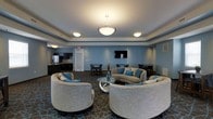 This single-floor common lounge is designed with both comfort and functionality in mind. The space is anchored by a sophisticated seating arrangement featuring curved, neutral-toned sofas and armchairs accented with teal throw pillows, all centered around a sleek glass coffee table. The combination creates an inviting conversation area well-suited for group gatherings or quiet relaxation.

The room's blue-grey walls lend a calm, contemporary ambiance, complemented by a tray ceiling with recessed lighting and a central pendant chandelier that provides warm, layered illumination throughout. A wall-mounted flat-screen television serves as a focal point on one wall, adding a multimedia element to the lounge.

Toward the back of the space, a compact kitchenette equipped with dark cabinetry, a sink, and a refrigerator offers convenient refreshment access. Additional seating is available at nearby café-style tables and chairs, providing flexible options for dining or casual use.

The richly patterned carpet featuring geometric motifs in deep tones grounds the space and adds visual interest underfoot, tying together the room's cohesive, modern aesthetic.