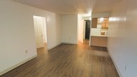 This single-floor residence offers a clean, functional layout with a welcoming open-concept living area as its centerpiece. Rich, dark hardwood-style plank flooring runs throughout the main space, adding warmth and a sense of continuity to the interior. The living area is bright and open, with neutral white walls that create a versatile canvas for any décor style. A textured ceiling adds a subtle architectural detail overhead.

The kitchen is seamlessly integrated into the living space via a raised peninsula counter, providing a natural division between the two areas while maintaining an open feel. Warm wood-toned upper cabinetry lines the kitchen walls, complemented by tile backsplash detailing and under-cabinet lighting that enhances visibility and ambiance. A refrigerator is visible within the kitchen, indicating a fully equipped cooking space.

From the main living area, two doorways lead to additional rooms, suggesting a multi-room floor plan with private spaces accessible from the central hub. One doorway opens to a hallway with continued hardwood-style flooring, while another leads to a darker interior room. The overall layout is practical and well-connected, making efficient use of the available space while maintaining a cohesive and comfortable atmosphere throughout.