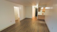 This single-floor residence offers a clean, functional layout with a welcoming open-concept living area as its centerpiece. Rich, dark hardwood-style plank flooring runs throughout the main space, adding warmth and a sense of continuity to the interior. The living area is bright and open, with neutral white walls that create a versatile canvas for any décor style. A textured ceiling adds a subtle architectural detail overhead.

The kitchen is seamlessly integrated into the living space via a raised peninsula counter, providing a natural division between the two areas while maintaining an open feel. Warm wood-toned upper cabinetry lines the kitchen walls, complemented by tile backsplash detailing and under-cabinet lighting that enhances visibility and ambiance. A refrigerator is visible within the kitchen, indicating a fully equipped cooking space.

From the main living area, two doorways lead to additional rooms, suggesting a multi-room floor plan with private spaces accessible from the central hub. One doorway opens to a hallway with continued hardwood-style flooring, while another leads to a darker interior room. The overall layout is practical and well-connected, making efficient use of the available space while maintaining a cohesive and comfortable atmosphere throughout.