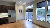 This well-appointed single-floor space showcases a thoughtfully designed open layout that seamlessly blends the kitchen and living areas. The interior is defined by warm hardwood flooring that runs throughout, contributing to an inviting and cohesive atmosphere.

The kitchen area is equipped with stainless steel appliances, including a full-sized refrigerator, and is complemented by dark cabinetry that provides a striking contrast against the light-toned walls. Modern track lighting along the ceiling ensures ample illumination across the space.

One of the most notable architectural features is the impressive ceiling height, which lends an airy, expansive feel to the interior. Large black-framed windows are positioned at an elevated level, drawing in abundant natural light while framing views of the surrounding tree canopy. A sliding glass door opens directly onto an outdoor deck fitted with metal railings, offering a seamless indoor-outdoor connection and a serene, nature-filled backdrop.

The overall ambiance of the space is contemporary and calm, with a clean, minimalist aesthetic enhanced by neutral wall tones and natural materials. The combination of elevated windows, generous ceiling height, and direct outdoor access makes this a bright and versatile space throughout the day.