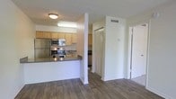 This single-floor residence offers a thoughtfully arranged layout designed for comfortable everyday living. The kitchen serves as a central feature of the space, outfitted with stainless steel appliances including a refrigerator, microwave, and electric range. Light wood cabinetry provides ample storage, while dark countertops offer a clean contrast. A raised breakfast bar peninsula defines the boundary between the kitchen and the adjoining living area, creating an open and connected feel throughout.

The flooring throughout the main living area is finished in rich, dark hardwood-style planks, lending warmth and a polished aesthetic to the interior. The walls are painted in a neutral, light tone that maximizes the sense of brightness and openness. Textured ceilings and flush-mounted lighting fixtures complete the overhead details.

A hallway leading from the main living area provides access to additional rooms, with a louvered bifold closet door offering convenient storage. The corridor is well-proportioned and connects the living spaces efficiently. HVAC vents are integrated into the walls, ensuring year-round climate comfort. Overall, the unit presents a clean, functional, and welcoming environment suitable for a range of residents.