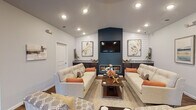 Located at 7405 Fegenbush Lane in Louisville, Kentucky, this single-floor community space is thoughtfully designed to offer both comfort and functionality. The interior showcases a cohesive modern aesthetic, anchored by rich hardwood-style flooring that runs throughout the open layout.

The central lounge area is the focal point of the space, featuring vaulted ceilings with recessed lighting that accentuate the dramatic accent wall in deep teal. A linear fireplace is framed beneath a wall-mounted flat-screen television, flanked by shiplap paneling and framed botanical artwork. Dark-stained built-in cabinetry with granite countertops lines both sides of the feature wall, adding refined storage and display space. Plush neutral sofas with warm orange accent pillows and a rustic wood coffee table complete the inviting seating arrangement.

An adjacent area flows naturally from the lounge, offering flexible seating with bistro-style tables and chairs positioned near natural light from exterior-facing windows and a glass door. This area provides a bright, casual gathering space.

A kitchenette is accessible from the main lounge, equipped with dark cabinetry, granite countertops, a sink, and a full-size refrigerator. A separate office nook with a ceiling fan adds practical utility to the overall layout.