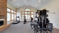 Located at 4600 Emerald Lakes Blvd in Powell, Ohio, this single-floor fitness center is designed to offer a well-rounded workout experience within an architecturally refined setting. The space is defined by soaring vaulted ceilings that create an open, airy atmosphere, while an impressive arched window arrangement along the front wall floods the interior with natural light and provides views of the surrounding greenery. Warm wood-trimmed windows are featured throughout, lending a classic and inviting aesthetic. A striking full-height brick fireplace serves as a prominent architectural focal point, complementing the neutral tones of the carpeted floors and light-colored walls. The main workout area is generously proportioned, accommodating a comprehensive range of equipment including treadmills, an elliptical trainer, a stationary bike, a multi-station cable weight machine, a rowing machine, and a full dumbbell rack with mirrors. French doors with glass panels open to the outdoors, providing both ventilation and easy access. Recessed lighting supplements the abundant natural light, ensuring the space remains well-lit at all times. A small adjoining area off the main floor provides additional functional space. The overall ambiance balances the energy of a professional fitness facility with the warmth of thoughtful architectural design.