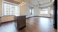 Located at 221–243 Bagley St in the heart of downtown Detroit, this single-floor unit blends industrial-loft character with modern finishes throughout. Rich hardwood flooring runs continuously across the open-plan living and kitchen areas, complemented by soaring exposed beam ceilings with visible structural elements that lend an authentic urban aesthetic.

The kitchen is a standout feature, outfitted with white shaker cabinetry, stainless steel appliances including a full-size refrigerator, microwave, and range, as well as a substantial dark-toned island with a concrete-style finish. Edison-bulb pendant lighting hangs above, adding warmth to the space. The open layout allows the kitchen to flow seamlessly into the expansive living area, where multiple large windows frame striking views of Detroit's downtown skyline and active streetscape.

The bedroom offers generous proportions with hardwood floors, cool-toned walls, and direct access to the bathroom. The bathroom is finished with large-format neutral tile flooring, a walk-in shower with built-in shelving, a wood-toned vanity with a white countertop, and a frameless mirror. Bold dark trim on doors and baseboards provides a sharp contrast throughout the unit, reinforcing the contemporary industrial design language that defines this well-appointed Detroit address.