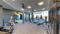 This fitness center is a thoughtfully designed, single-floor facility offering a comprehensive workout environment. The main exercise area is anchored by a row of cardio machines — including multiple treadmills, ellipticals, and stationary bikes — each equipped with individual display screens and positioned to take full advantage of the floor-to-ceiling windows along one wall. These expansive windows flood the space with natural light and provide open outdoor views, creating an energizing atmosphere.

The strength training zone is well-appointed, featuring a tiered dumbbell rack with a full range of free weights, a weight bench, and a multi-function cable machine for resistance training. A rack of medicine balls and stability balls in vibrant colors adds a functional training element to the space. Patterned, low-pile carpeting covers the floors throughout, providing comfort underfoot while complementing the cool-toned gray and blue wall palette.

A secondary studio area, accessible through glass-paned interior doors, offers additional open floor space suited for stretching, yoga, or group exercise. This room features built-in shelving, a ceiling fan, and a recessed tray ceiling detail. Combined recessed and panel lighting ensures consistent illumination throughout both areas, rounding out a polished and functional fitness environment.