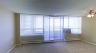 This single-floor space is defined by its striking wall of floor-to-ceiling windows dressed with vertical blinds, allowing for adjustable natural light throughout the day. A sliding glass door opens directly onto a balcony, offering sweeping outdoor views that appear to extend toward a distant horizon — a standout feature that gives the interior an airy, open feel. The main living area is finished with neutral wall-to-wall carpeting that complements the light, understated color palette of the space. Baseboard heating units run along the base of the window wall, providing efficient climate control, while a window-mounted air conditioning unit ensures comfort during warmer months. The smooth, low-profile ceiling contributes to a clean, uncluttered aesthetic. The open layout provides a versatile canvas, well-suited for a variety of furniture arrangements and uses. The abundance of glazing ensures the space remains bright and connected to the outdoors, making natural light a defining characteristic of the overall ambiance. The combination of practical climate control features and panoramic views makes this a functional yet visually engaging single-level space.