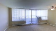 This single-floor space is defined by its striking wall of floor-to-ceiling windows dressed with vertical blinds, allowing for adjustable natural light throughout the day. A sliding glass door opens directly onto a balcony, offering sweeping outdoor views that appear to extend toward a distant horizon — a standout feature that gives the interior an airy, open feel. The main living area is finished with neutral wall-to-wall carpeting that complements the light, understated color palette of the space. Baseboard heating units run along the base of the window wall, providing efficient climate control, while a window-mounted air conditioning unit ensures comfort during warmer months. The smooth, low-profile ceiling contributes to a clean, uncluttered aesthetic. The open layout provides a versatile canvas, well-suited for a variety of furniture arrangements and uses. The abundance of glazing ensures the space remains bright and connected to the outdoors, making natural light a defining characteristic of the overall ambiance. The combination of practical climate control features and panoramic views makes this a functional yet visually engaging single-level space.