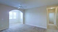 This single-story space presents a clean, neutral aesthetic with wall-to-wall carpeting and light beige walls that create a warm, understated ambiance. The main living area is highlighted by a ceiling fan that adds both functionality and a subtle decorative touch. A standout architectural feature is the rounded arched passageway that leads into an adjoining area, lending the space a classic, refined character. This secondary area benefits from natural light streaming through two windows, brightening the interior and creating an inviting alcove-like feel. White baseboards run throughout, providing a crisp contrast to the soft carpet and walls. The overall layout feels open and airy, offering a versatile blank canvas suitable for a variety of furnishing arrangements. The consistent use of neutral tones across all surfaces contributes to a cohesive and calm atmosphere throughout the space.