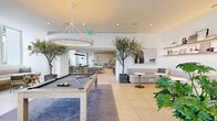 Located at 2000 Biscayne Blvd in Miami's vibrant Edgewater neighborhood, this generously proportioned amenity space spans a single floor and offers a thoughtfully designed environment for socializing and recreation. The open-plan layout is anchored by large-format porcelain tile flooring and crisp white walls, creating a bright, modern aesthetic throughout.

The game room is a standout feature, centered around a substantial pool table beneath a sculptural circular pendant light, and complemented by a full-length shuffleboard table positioned against a wall-mounted flat-screen TV. Mature olive and tropical trees add organic warmth to the space, while a wood-slat accent wall and warm timber ceiling details introduce natural texture.

A well-equipped social kitchen features a dark-topped island with bar seating, pendant lighting, and built-in cabinetry, ideal for community gatherings. Adjacent dining and lounge areas offer a mix of banquette seating, pedestal tables, and casual chairs. A curved upholstered banquette with floating display shelves provides an intimate nook for conversation.

Floor-to-ceiling windows and glazed doors flood the interior with natural light and frame views of Miami's skyline and swaying palm trees. A bright corridor connects the various zones, leading to an outdoor terrace area, further enhancing the seamless indoor-outdoor living experience.