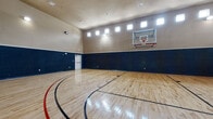 This remarkable indoor sports facility centers around a full-size basketball court with a gleaming, polished hardwood floor finished with regulation black and red court markings. Soaring double-height ceilings give the space an expansive, open-air feel, while rows of clerestory windows along the upper walls flood the interior with abundant natural light throughout the day. The walls are finished in a two-tone design — a deep navy lower half paired with a warm taupe upper section — lending the court a clean, professional aesthetic. A glass-backboard basketball hoop is mounted at one end, and recessed and surface-mounted lighting ensures excellent visibility for evening play. One side of the court features large interior windows that look directly into a well-equipped fitness room, keeping the athletic spaces visually connected. French doors on another wall open to an adjacent covered outdoor area complete with a fireplace and television, perfect for gathering after a game. An elegant wrought iron balcony overlooks the court from above, adding an architectural focal point. Additional doors provide convenient access to supporting rooms. Together, these elements create a cohesive, high-caliber athletic and entertainment environment designed for serious recreation and active living.
