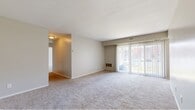 This single-floor residence offers a functional and comfortable layout with four bedrooms, two bathrooms, a living room, kitchen, hallway, and closet. The living room is a generously sized, open area featuring soft wall-to-wall carpeting, neutral beige tones, and large sliding glass doors fitted with vertical blinds that allow an abundance of natural light and provide access to an outdoor view. A built-in heating unit is tucked beneath the slider, adding a practical touch to the space. The galley-style kitchen is equipped with dual-tone gray cabinetry lining both walls, a gas range, a dishwasher, a sink, and warm hardwood-style flooring that contrasts nicely with the light countertops. A louvered closet door adjacent to the kitchen provides convenient storage access. The four bedrooms are carpeted with the same neutral palette as the living room, each offering a calm and restful atmosphere. The primary bedroom is comfortably sized, featuring a window for natural light. The two bathrooms include one with a white vanity, framed mirror, and light wood-style flooring, and another with a full white-tiled bathtub and shower surround. A connecting hallway ties all rooms together in a cohesive, easy-to-navigate floor plan.